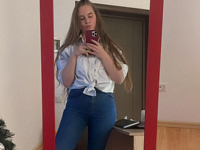 Dooris with long brown hair, white button-up shirt tied at the front, blue high-waisted jeans, holding a red phone, standing in front of a mirror, reflecting her image.