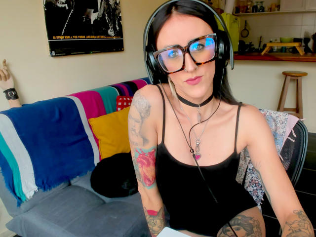 Envyv, com cabelo preto, óculos e fones de ouvido, vestindo uma regata preta, mostrando tatuagens nos braços, sentada em um sofá com um cobertor colorido e um colar..