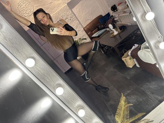 MisissFire in black shorts, sheer black stockings, and boots, stretching in front of a mirror with lights. Long brown hair, phone in hand, showing off her ass.