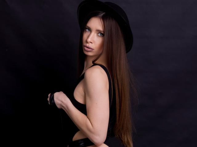 MisissFire with long brown hair, wearing a black hat and black top, looking back with blue eyes, holding black straps, showing her tits and a sexy, confident face.