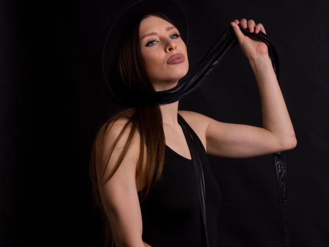 MisissFire with long brown hair, wearing a black hat and sleeveless top, holding black fabric over her head, puckering her lips. She's got a cute ass and pale skin.
