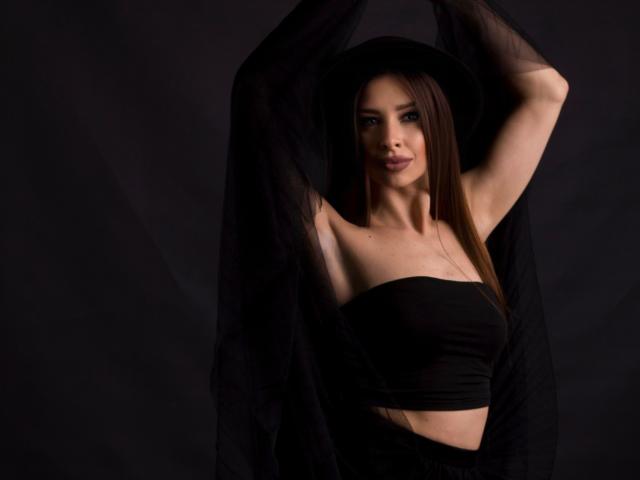 MisissFire with long brown hair, wearing a black hat and strapless top, holding a black sheer cloth above her head, showing her small tits and a slight smile.