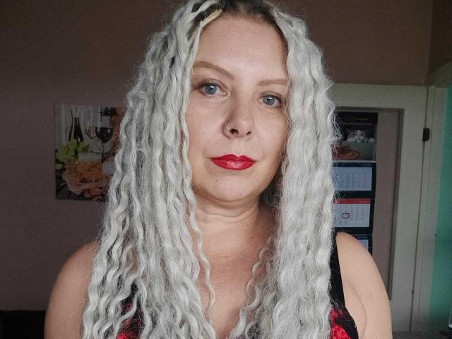LuxuryWhite, met witte dreadlocks, rode lippenstift, een neusring en blauwe ogen, draagt een zwart-rode top met bloemenprint en heeft een zacht, licht mollig gezicht en een lichte huid..