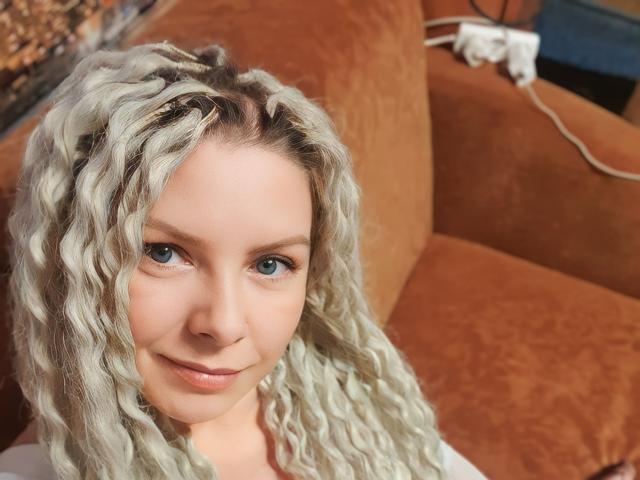 LuxuryWhite with curly white hair, blue eyes, and a slight smile, sitting on an orange-brown couch. Plug in the wall socket, white cord visible.