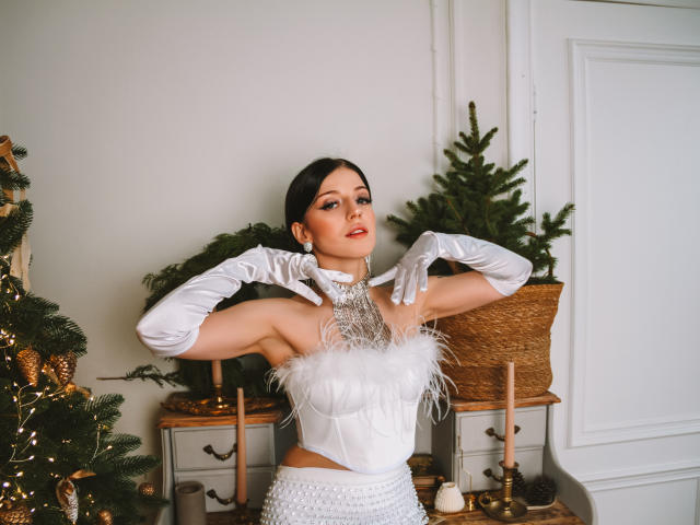 MiaFane, vêtue d'un haut blanc et duveteux sans bretelles, d'une jupe blanche à taille haute et de longues gants blancs, les mains levées, avec des cheveux foncés, du rouge à lèvres rouge et un collier en argent, se tient devant un sapin de Noël..