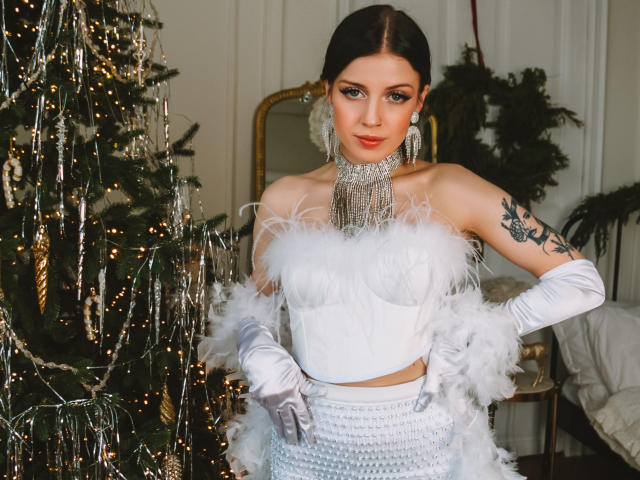 MiaFane, avec un haut blanc en plumes, des gants argentés et une jupe taille haute, se tient près d'un sapin de Noël décoré, et elle a un tatouage sur son bras droit et des yeux bleus..