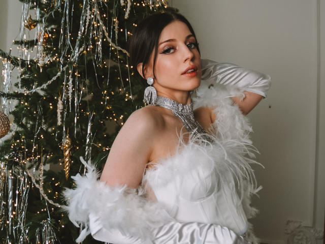 MiaFane, vêtue d'une robe blanche avec des plumes, un collier en argent et des boucles d'oreilles, se tient devant un sapin de Noël décoré, portant de longs gants blancs, et a l'air séduisante, montrant sa poitrine et ses fesses..