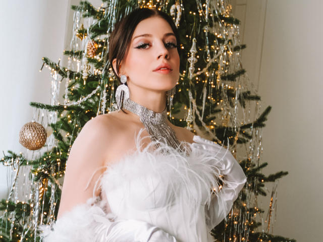 MiaFane, vêtue d'une robe blanche avec des plumes, un collier en argent et des boucles d'oreilles, se tient devant un sapin de Noël décoré de boules dorées et de guirlandes lumineuses, affichant une attitude confiante, les mains près de sa poitrine..