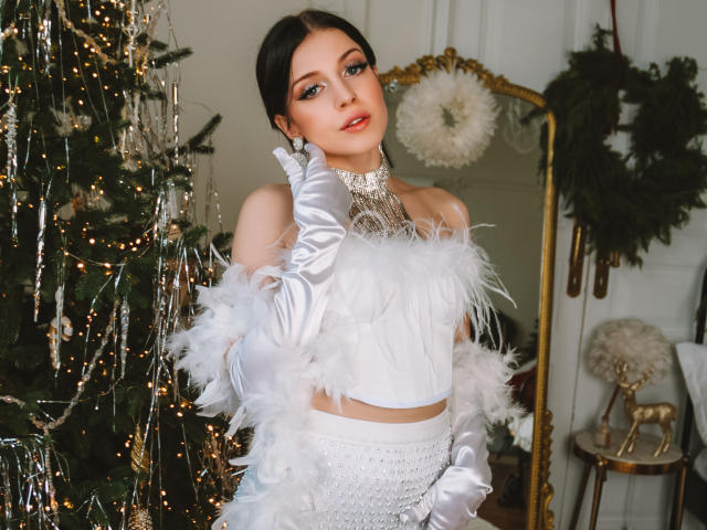MiaFane, vêtue d'une robe blanche avec des plumes, des gants et un collier, se tient près du sapin de Noël, regardant l'appareil photo, montrant ses seins, dans une pièce élégante avec un miroir orné et une couronne..