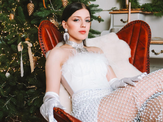 MiaFane, vêtue d'une robe blanche ornée de plumes, avec des boucles d'oreilles argentées et des bas en dentelle blanche, assise sur un fauteuil en velours rouge, portant des gants blancs, à côté d'un sapin de Noël décoré..