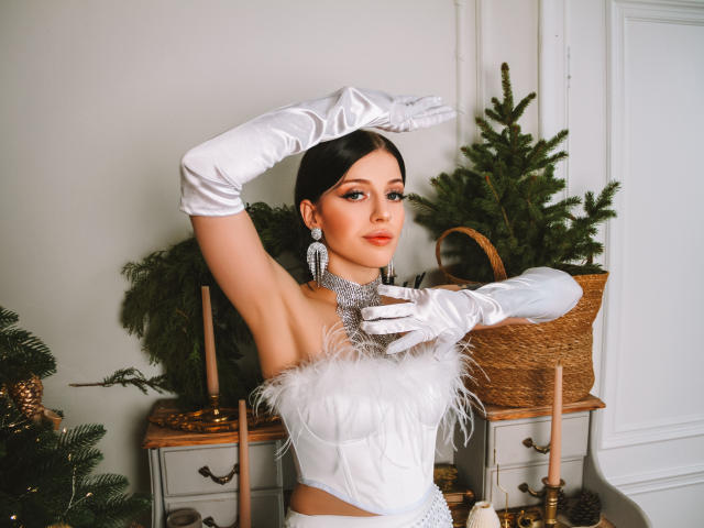 Mia Fane, avec ses cheveux foncés, portant un corset blanc orné de plumes, de longs gants blancs et des boucles d'oreilles argentées, touchant son menton, regardant l'appareil photo, avec des sapins verts et des bougies en arrière-plan..