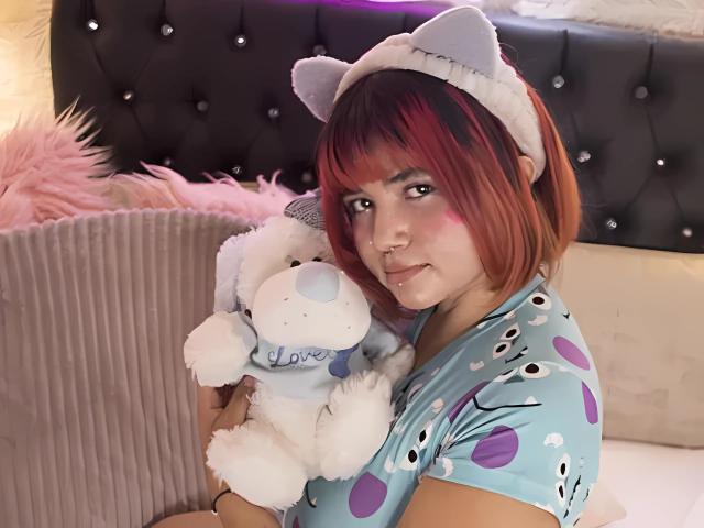 NicolRitz with red-brown bob, wearing cat ears, blue polka-dot pajamas, holding white stuffed dog, pink blush on cheeks, sitting on bed, smiling, visible nose piercing.