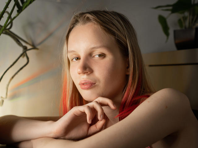 Beatrica Klein, con cabello rubio y teñido de rojo, piel clara y senos pequeños, sentada con los brazos cruzados, mirando a la cámara, luz suave, labios fruncidos, ligera sonrisa, ojos verdes..