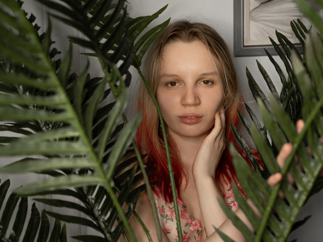 BeatricaKlein with red-tipped blonde hair, floral dress, and pale skin, standing behind green palm leaves, holding her cheek, looking at camera with a neutral expression.