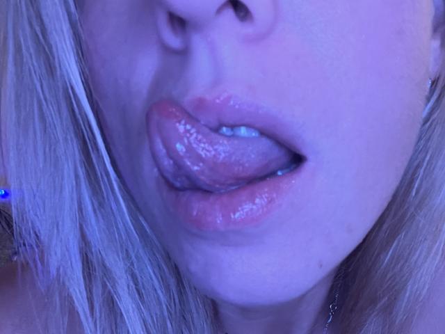 SandraSwweet's glossy pink lips are slightly parted with her tongue out, showing a wet, shiny surface under blue-purple lighting. Her blonde hair is partially visible.