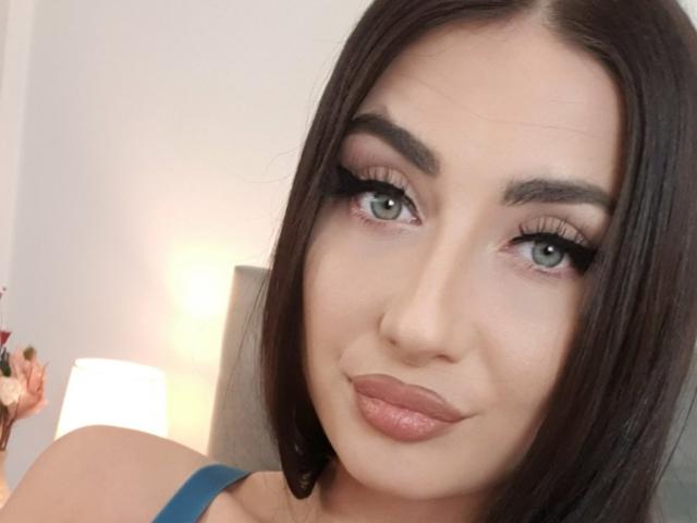 A DreamsGirl tem cabelo castanho escuro, olhos verdes e lábios rosados cheios, com cílios pretos grossos. Ela está vestindo uma blusa azul e está olhando para a câmera com um leve sorriso..