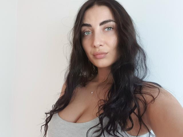 Ragazza con capelli neri ondulati, occhi azzurri e un sorriso carino, che indossa una canotta grigia che mette in mostra il suo seno prosperoso e una piccola collana, e che guarda verso la fotocamera..