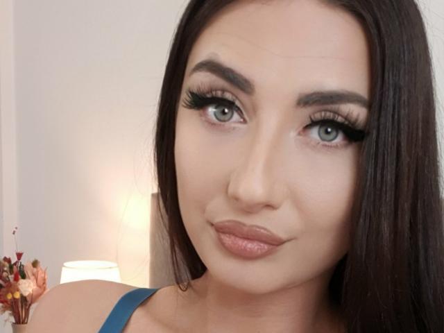 DreamsGirl a de longs cheveux bruns foncés, des yeux verts et des lèvres pulpeuses avec un rouge à lèvres rose brillant. Elle porte des bretelles bleues et a l'air séduisante avec de longs cils noirs..