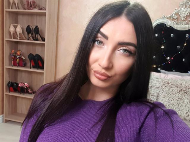 Una ragazza con lunghi capelli neri, che indossa un maglione viola, ha labbra rosa carnose e occhi verdi, ed è in piedi in una stanza con una mensola di tacchi alti..