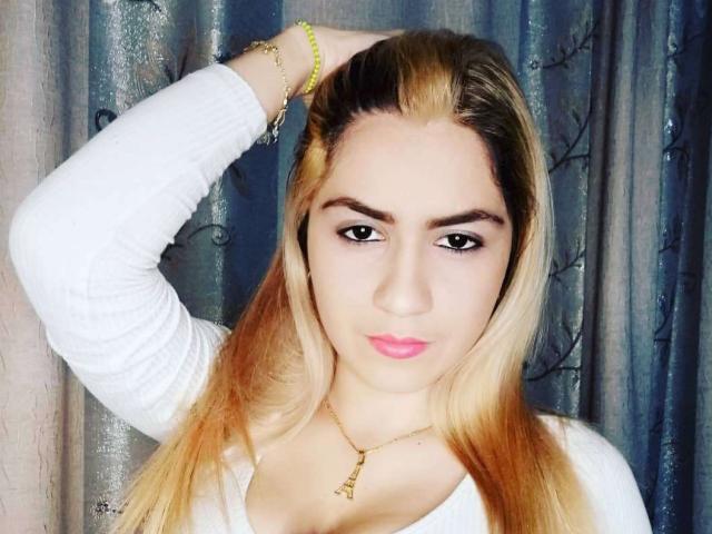 Steven e Paty, com cabelo loiro, camisa branca de manga comprida, batom rosa, mão no cabelo, pulseira amarela, colar com a Torre Eiffel, expressão séria, seios médios, pele clara, olhos azuis e corpo esguio..