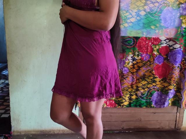 Murielles trägt ein lila Spitzenkleid, wobei ihr Hintern leicht zu sehen ist. Sie steht mit einem Bein angewinkelt, ein Arm über der Brust verschränkt, und ihr langes, braunes Haar ist sichtbar..