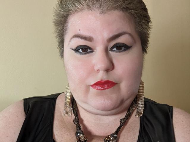 KaryQueen has short brown hair, black eyeliner, red lipstick, and wears a black leather top with gold and black bead necklace. Her face is slightly sweaty.
