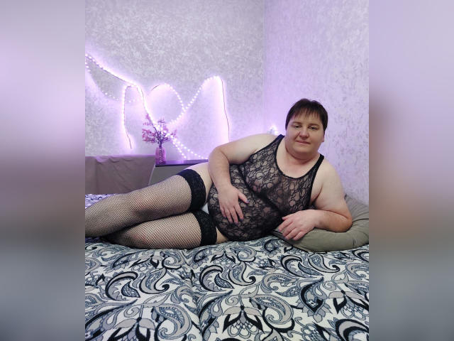 LimonkaFire, plus-sized with short brown hair, lies on a bed wearing a black lace lingerie top, black fishnet stockings, and smiling. Her hand rests on her belly.