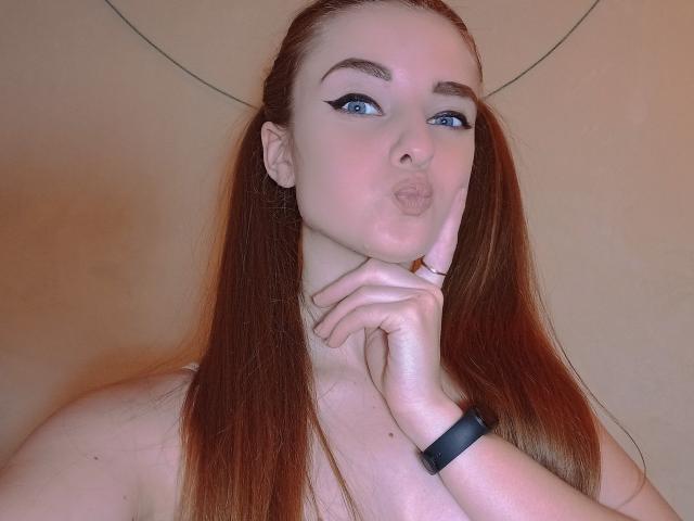 KariMillie with pouty lips, blue eyes, and red pigtails, wearing a black wristband, her hand on her neck, exposing her bare shoulders and tits.