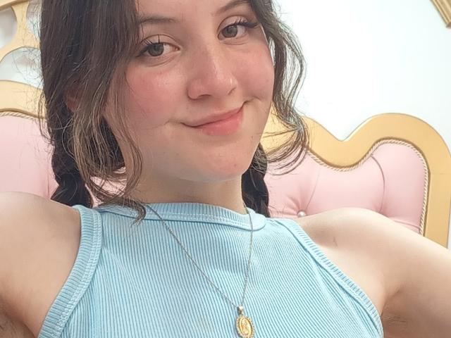 ChloeDulce met een schattende glimlach, gekleed in een blauw geribbeld tanktop, waarbij haar blote schouders en armen zichtbaar zijn, met een gouden ketting en gevlochten bruin haar..