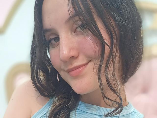 ChloeDulce a les cheveux longs, bruns foncés, un joli sourire et une peau claire. Elle porte un haut bleu, côtelé et sans manches, qui met en valeur sa petite poitrine ferme..