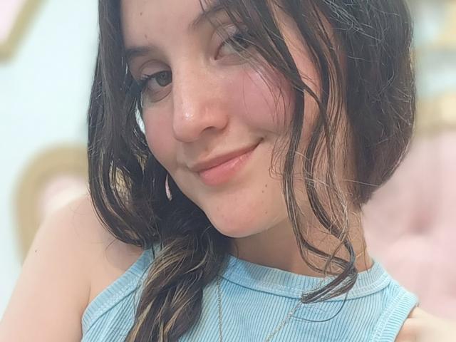 ChloeDulce hat feuchtes, braunes Haar, ein süßes Lächeln und helle, rosafarbene Wangen. Sie trägt ein blaues, geripptes Tanktop, das ihre kleinen, prallen Brüste zeigt..