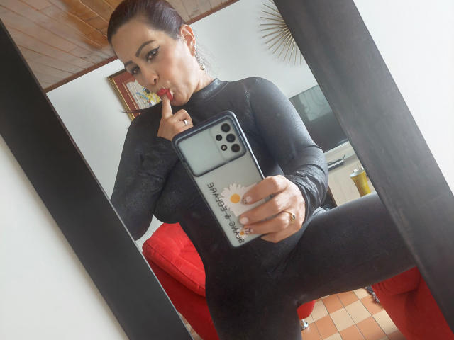 MelissaDelici met donker haar in een knot, gekleed in een zwarte lange mouwen top, met een telefoon in haar hand, haar vinger in haar mond, een spiegelzelfportret, waarbij haar achterwerk te zien is, een rode bank op de achtergrond..