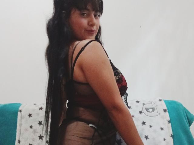 Lana Lorens, con el pelo largo y negro, mirando hacia atrás, vestida con una lencería negra transparente, que deja ver sus pechos y glúteos, sentada sobre una manta con un estampado de estrellas azules y blancas..