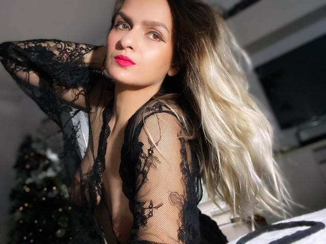 ReveMignoneX with blonde-to-brown ombre hair, black lace robe, pink lipstick, hand in hair, visible small tits, soft light, Christmas tree blurred in background.