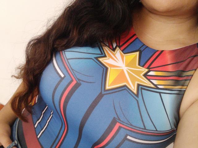 CamilaHot wearing a blue and red superhero-style top with a yellow star, her large, round tits prominently displayed, long wavy brown hair falling over her shoulder.