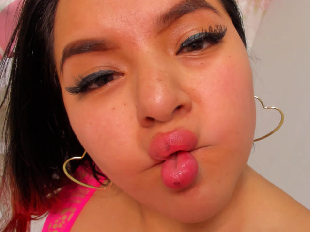 Jazmine Ferra, met volle, roze lippen, hartvormige oorbellen en donkere eyeliner, maakt een "eendensnot" gezicht, waarbij haar mollige gezicht en bruine wenkbrauwen zichtbaar zijn, met steil, zwart haar..