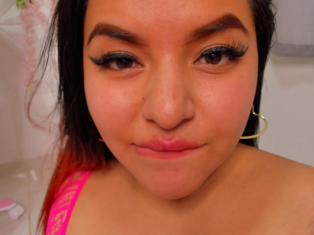 Le visage de Jazmine Ferra, avec ses sourcils épais, son eye-liner noir, son rouge à lèvres rose et un léger froncement de lèvres. Elle porte un haut rose et une boucle d'oreille dorée. Sa peau est bronzée..