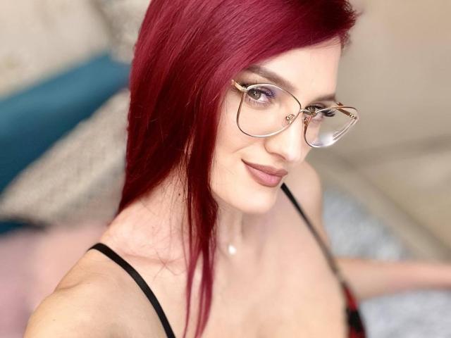 Rosalyn, con cabello rojo, usando gafas, y un top negro con tirantes, mostrando sus senos, sonriendo, con piel clara y un rostro lindo, en un selfie..