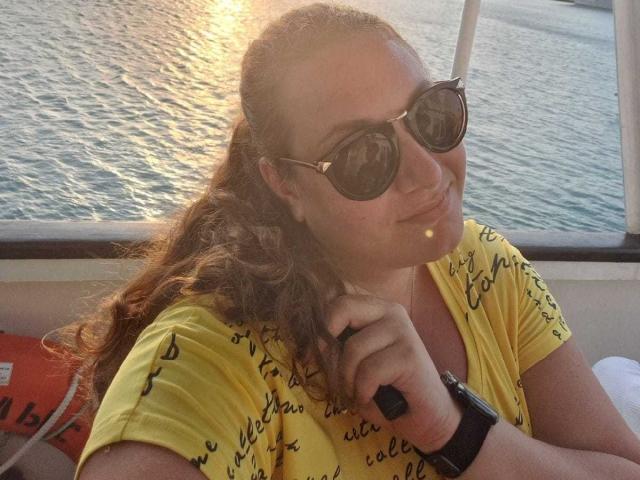 NickiAmabel, avec ses cheveux bruns bouclés, portant une chemise jaune, des lunettes de soleil noires et une montre noire, faisant la moue avec un petit reniflement sur sa lèvre supérieure, assise sur un bateau..