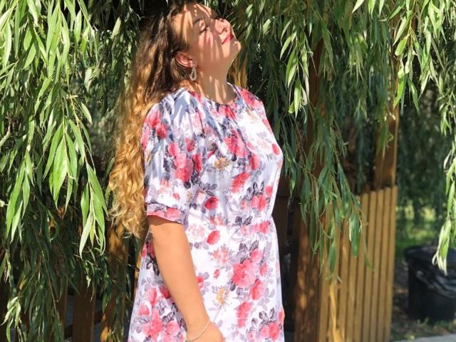 NickiAmabel with curly blonde hair, eyes closed, leaning against green weeping willow leaves, wearing a floral shirt, hands in pockets, sunlight on face, wood fence in background.