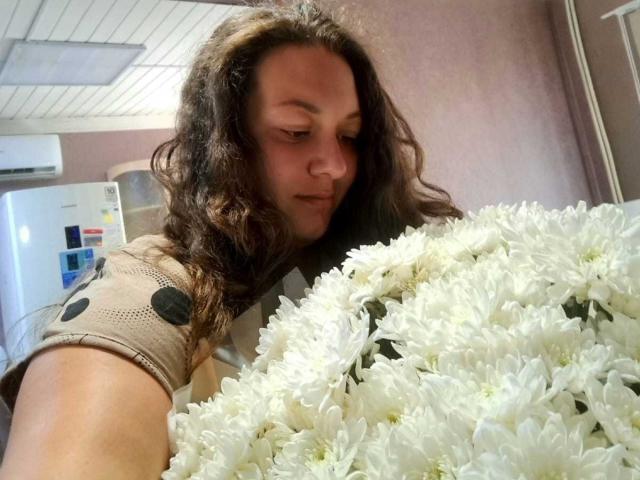 NickiAmabel, avec ses longs cheveux bruns ondulés, sa chemise beige et son bouquet de marguerites blanches, regarde vers le bas et tient les fleurs devant elle. Elle a une expression douce et adorable..