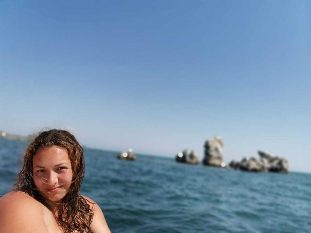NickiAmabel, con el cabello castaño mojado, sonriendo a la cámara, sin sostén, mostrando sus hombros desnudos y pequeños senos, de pie en el océano azul con rocas al fondo..