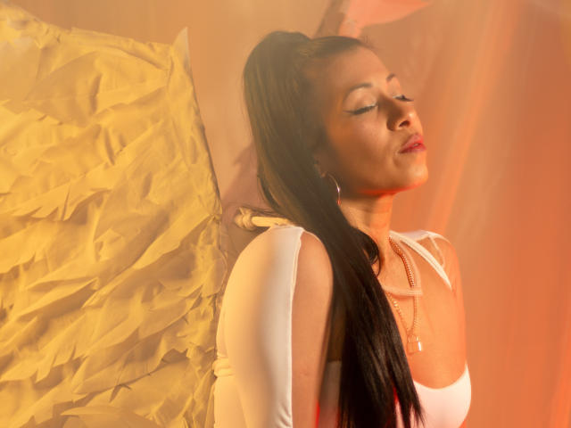 LaurenHills with long black hair, eyes closed, wearing white top, red lipstick, earrings, and necklace, leaning against crumpled yellow paper, looking relaxed, with soft orange light.