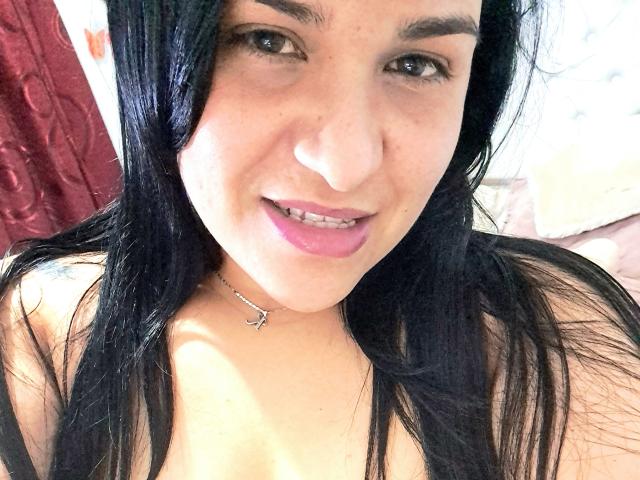 VivianaThompson has long black hair, dark eyebrows, pink lipstick, and a slight smile, showing her teeth. She's wearing a black top and a silver necklace with a small anchor.