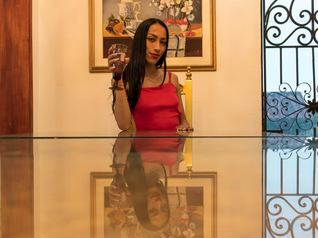 Thamarar with long black hair, red dress, holding glass, reflection on glass, ornate wall, gold frame, and black iron window. Boobs visible under dress, looking slightly down.