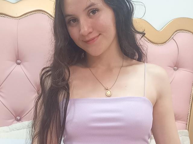 ChloeDulce mit langen, welligen braunen Haaren, lächelnd, trägt ein lila Top mit Spaghetti-Trägern, das ihre kleinen Brüste zeigt, und eine goldene Halskette mit einem runden Anhänger, sitzend im Bett..