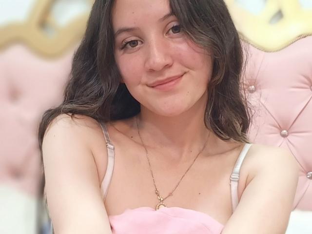 ChloeDulce, con cabello castaño ondulado, sonriendo suavemente, vestida con un top rosa con tirantes, un collar dorado, y un cabecero rosa con borlas en el fondo. Los senos parcialmente cubiertos por tela rosa..