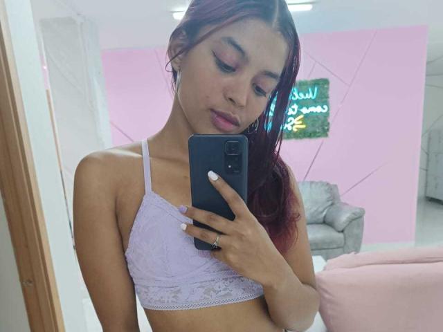 HilaryCook, avec des cheveux roux, portant un soutien-gorge en dentelle lilas, prenant un selfie devant un miroir, montrant ses seins, tenant un téléphone bleu, avec des paupières roses et des ongles blancs..