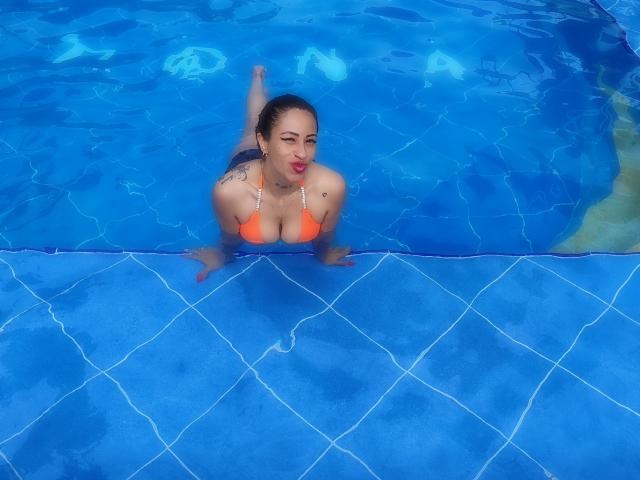 LunaCandy mit orangefarbenem Bikini-Oberteil, blauem Wasser und einem poolförmigen Boden mit Diamantmuster, kniend, wobei ihre Brüste sichtbar sind, Tätowierungen an ihrer rechten Schulter, roter Lippenstift, wellendes Wasser..