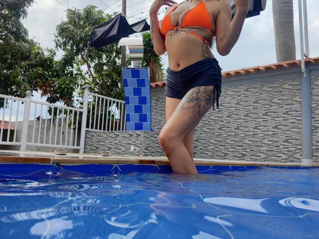 LunaCandy mit orangefarbenem Bikini-Oberteil, schwarzen Shorts, hält ein schwarzes Handtuch, steht in einem blau gefliesten Pool, hat ein Tattoo an der rechten Oberschenkelinnenseite, das Wasser reicht ihr bis zu den Knien, bewölkter Himmel darüber..